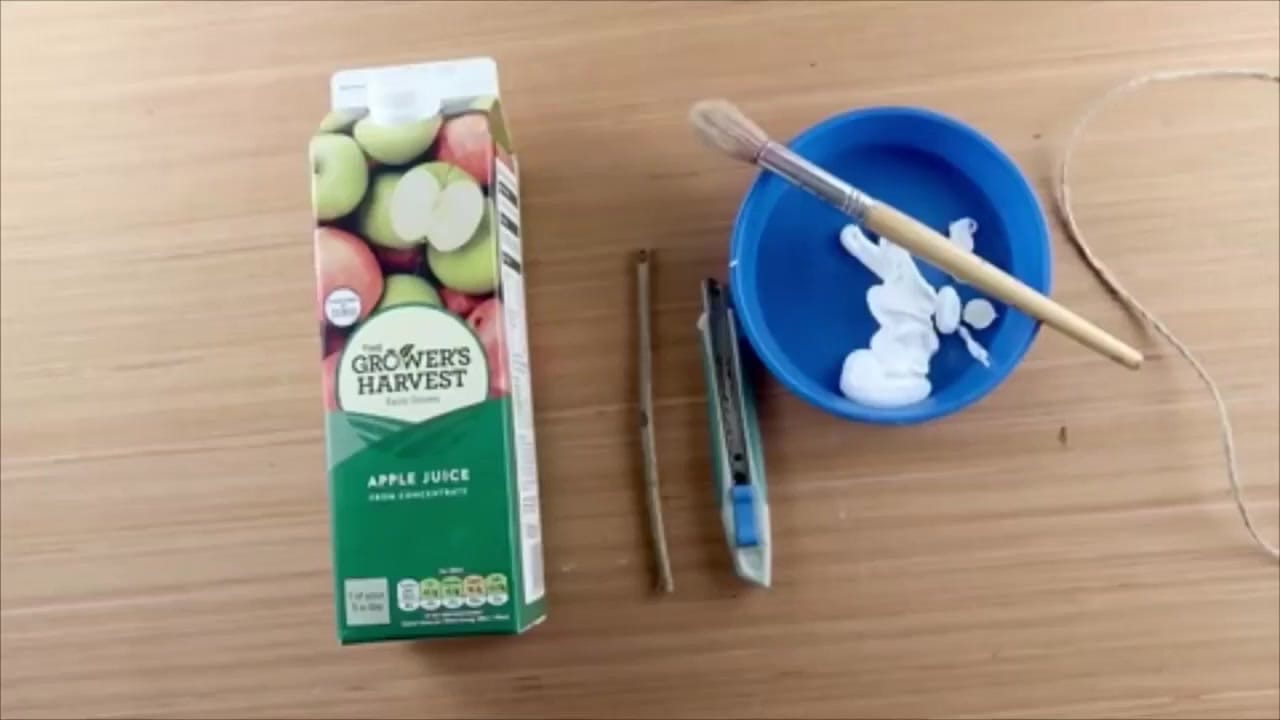 Craft materials laid out for a recycled bird feeder activity, including a clean juice carton, scissors, acrylic paint, decorations, a twig, and bird seed