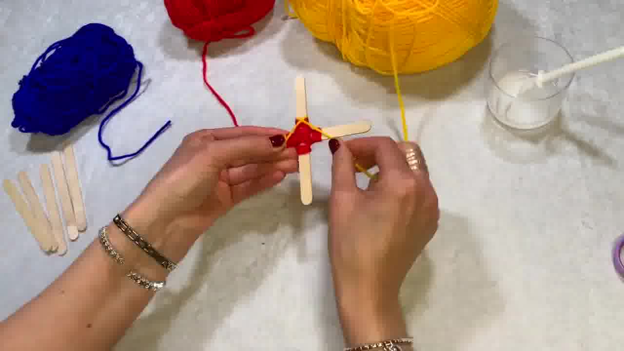 Two wooden lolly sticks glued together in a cross shape, ready to begin the Ojo de Dios craft. Coloured wool tied around the centre of the cross, starting to wrap around the middle.
